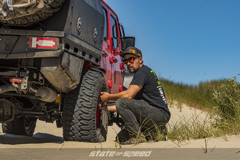 man checks the tire pressure on a red jeep gladiator on the beach