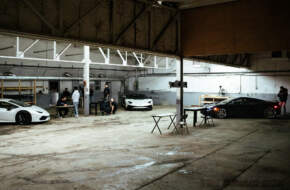The whole team working in big garage with two Lamborghinis and a McLaren parked inside